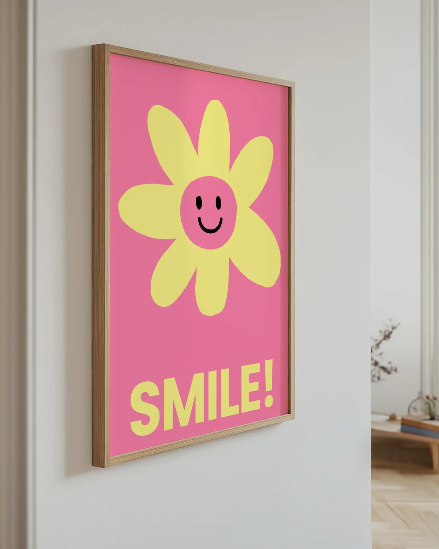 Framed artwork with a yellow flower and 'SMILE!' text on a pink background, hanging on a wall.